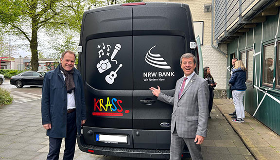 Stephan Keller, Oberbürgermeister der Stadt Düsseldorf, und Eckhard Forst, Vorstandsvorsitzender der NRW.BANK, stehen vor einem schwarzen Transporter mit dem Logos des Vereins Krass e. V. und der NRW.BANK.