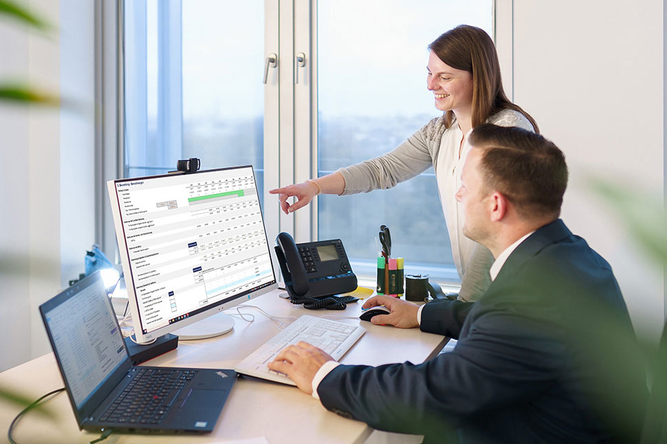 Auf dem Bild sieht man einen Unternehmer und eine Unternehmerin. Die Frau zeigt dem Mann eine Unternehmensbewertung auf dem Desktop. Sie wollen ein Unternehmen kaufen.