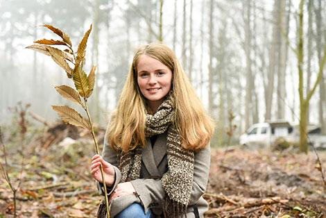 Eine junge Frau sitzt auf einem herbstlichen Waldboden und hält einen Ast mit Blättern in der Hand.