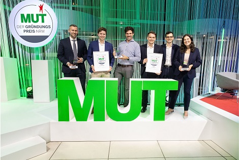 Sechs Personen stehen auf einer Bühne hinter großen grünen Buchstaben „MUT“ und halten Urkunden sowie Auszeichnungen; links oben ist das Logo „MUT – Der Gründungspreis NRW“ vor einem grün-blau beleuchteten Event-Hintergrund zu sehen.