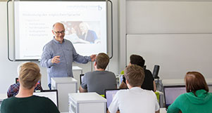 Ein Lehrer steht vor einer digitalen Tafel und unterrichtet eine Klasse Jugendlicher.