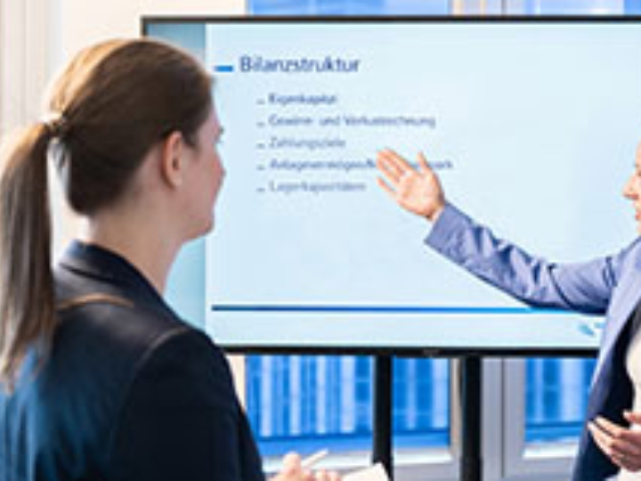 Eine Frau hält vor einer weiteren Frau und einem Mann eine Präsentation an einem Bildschirm