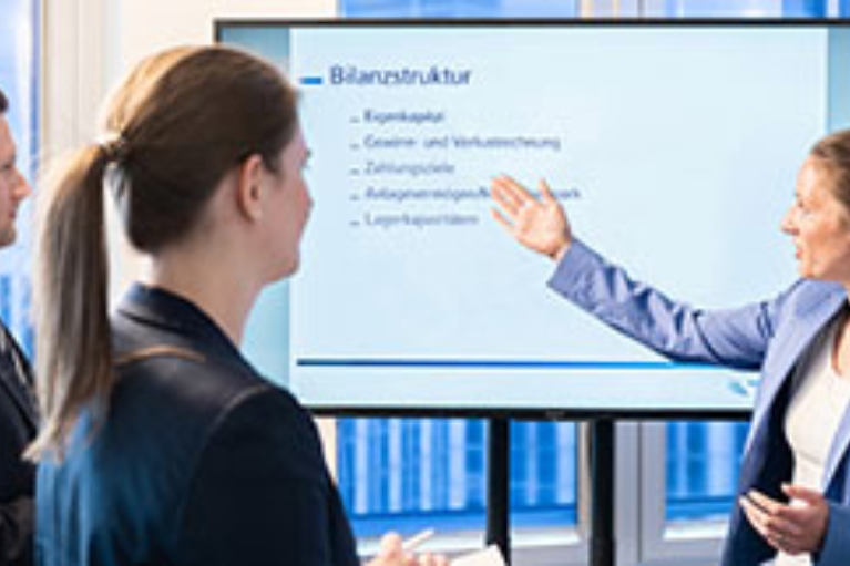 Eine Frau hält vor einer weiteren Frau und einem Mann eine Präsentation an einem Bildschirm