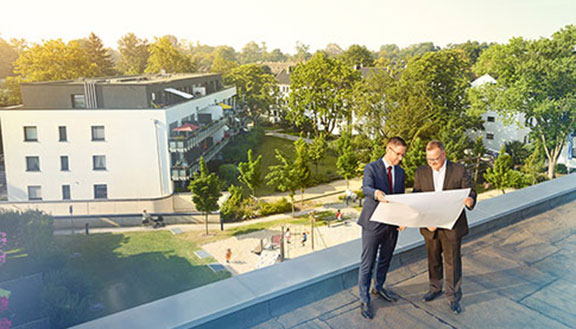 Zwei Männer stehen auf einem Hausdach und betrachten einen Bauplan. Im Hintergrund Bäume und ein Spielplatz