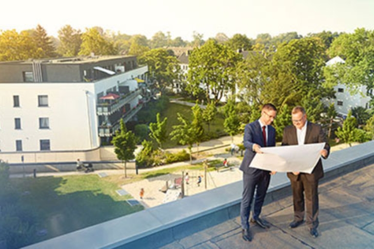 Zwei Männer stehen auf einem Hausdach und betrachten einen Bauplan. Im Hintergrund Bäume und ein Spielplatz