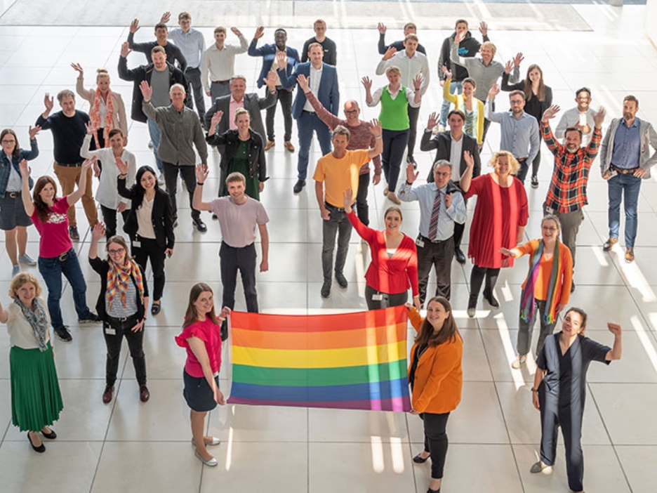Bunt gekleidete Mitarbeiterinnen und Mitarbeiter der NRW.BANK halten einen Regenbogen-Fahne hoch