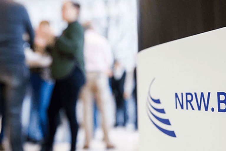 Verschwommen im Hintergrund stehen Menschen zusammen und unterhalten sich auf einer NRW.BANK-Veranstaltung.