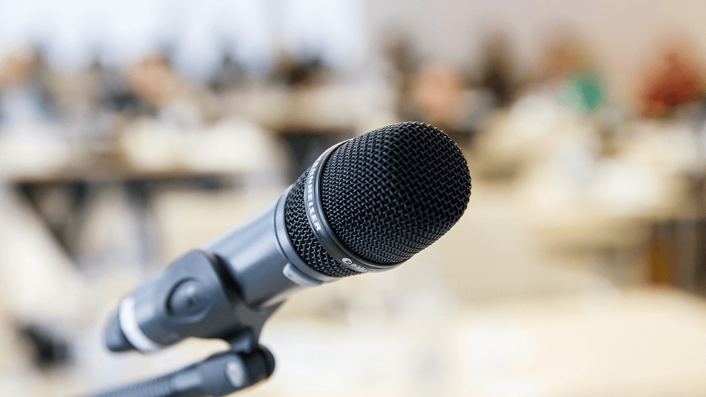 Ein Mikrofon auf einem Ständer, im Hintergrund unscharf sitzen Menschen an Tischen