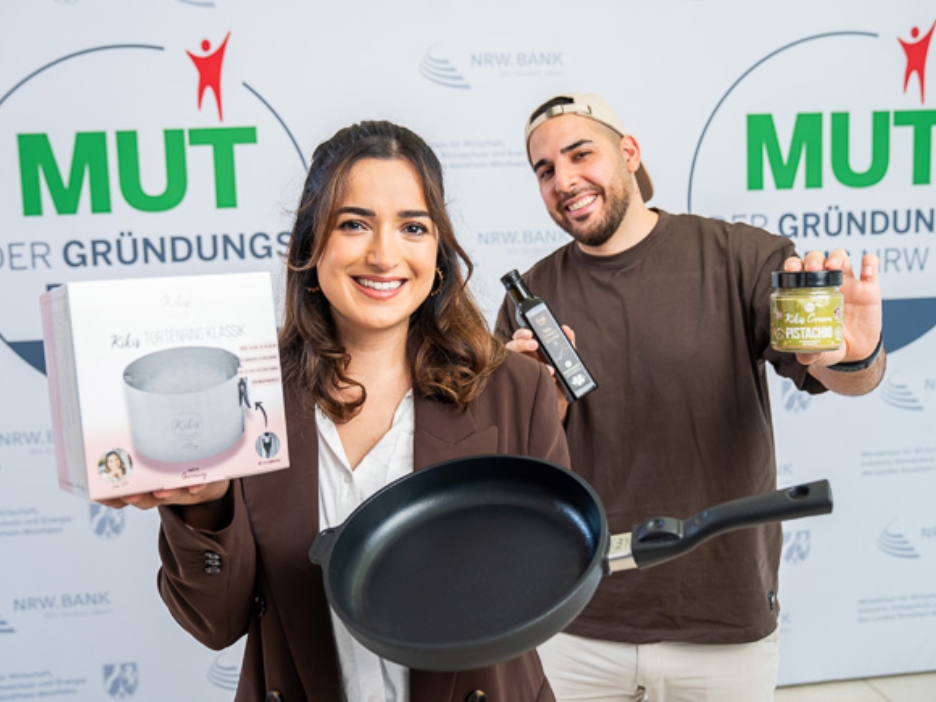 Kiki Aweimer und Hamza Aweimer von Kikis Kitchen vor dem MUT - DER GRÜNDUNGSPREIS NRW-Logo