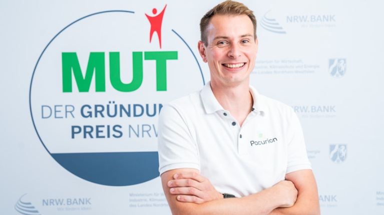 Dominik Leufgen von Pacurion vor dem MUT - DER GRÜNDUNGSPREIS NRW-Logo