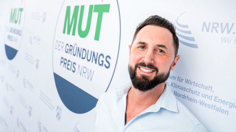 Robin Klein von Pader Solartechnik vor dem MUT - DER GRÜNDUNGSPREIS NRW-Logo