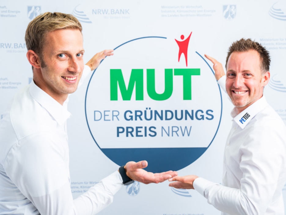 Christian Plett und Florian Eickelmann von PETec Table Lift Systems vor dem MUT - DER GRÜNDUNGSPREIS NRW-Logo