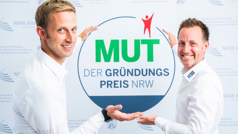 Christian Plett und Florian Eickelmann von PETec Table Lift Systems vor dem MUT - DER GRÜNDUNGSPREIS NRW-Logo