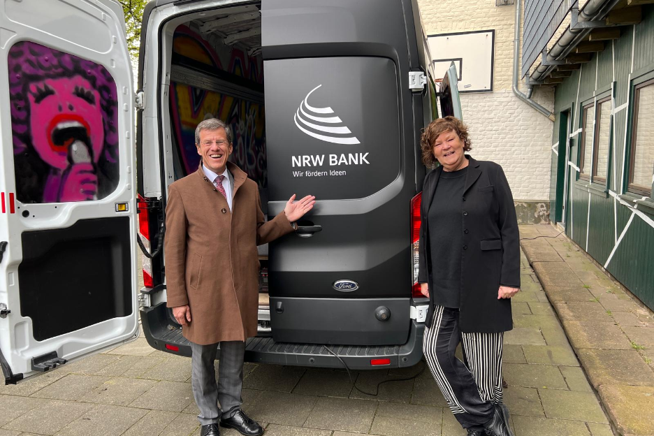 Das Foto zeigt einen Mann und eine Frau, die hinter einem mattschwarzen Sprinter stehen. Auf der linken Tür ist innen ein Graffiti zu sehen, auf der rechten Tür steht außen in weißer Schrift : NRW.BANK. Wir fördern Ideen.