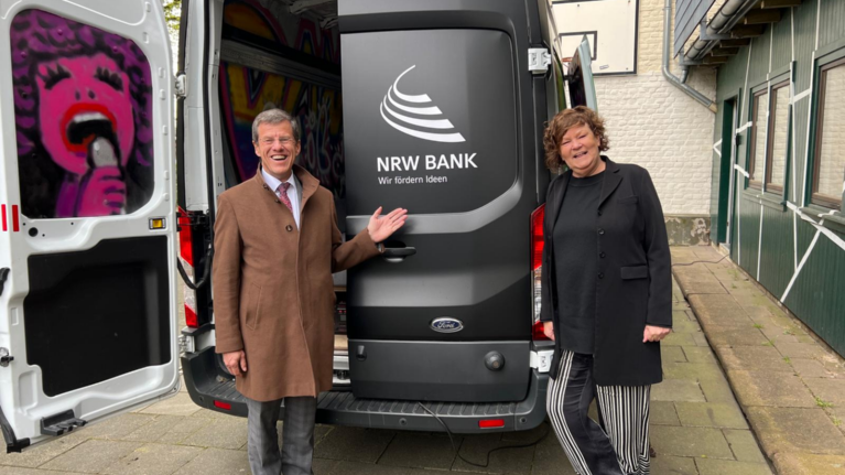 Das Foto zeigt einen Mann und eine Frau, die hinter einem mattschwarzen Sprinter stehen. Auf der linken Tür ist innen ein Graffiti zu sehen, auf der rechten Tür steht außen in weißer Schrift : NRW.BANK. Wir fördern Ideen.