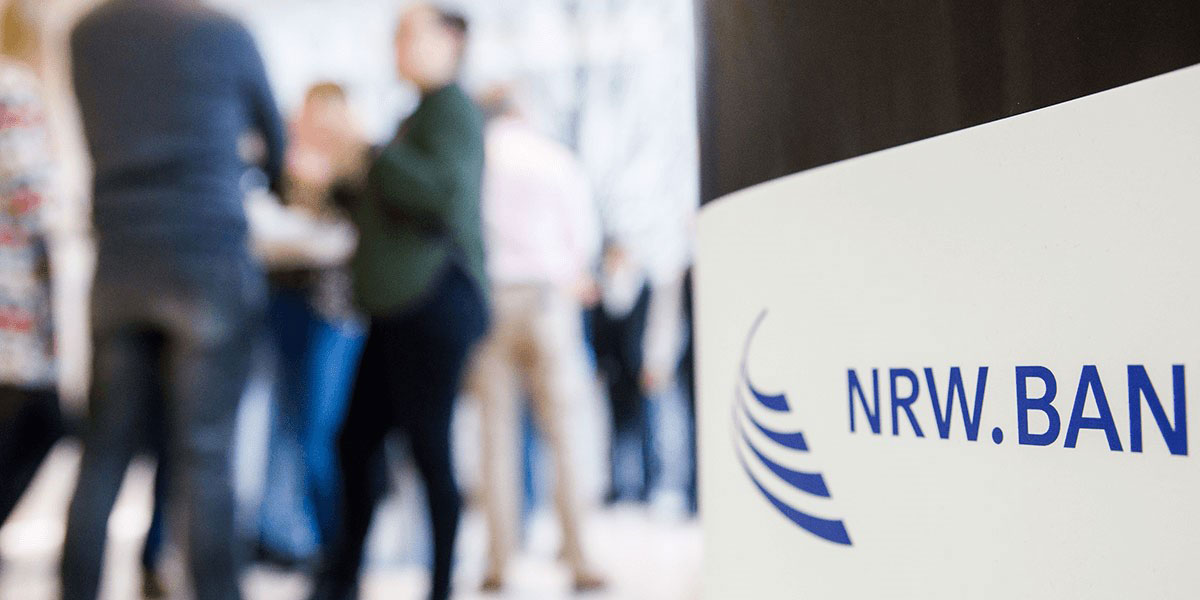 Im Vordergrund ein weißes Schild mit NRW.BANK-Logo, im Hintergrund unscharf eine Gruppe Menschen im Gespräch