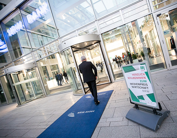 Ein Mann betritt das Foyer der NRW.BANK. Vor der Tür ein Schild: Herzlich willkommen zum MEDIENPREIS WIRTSCHAFT NRW 2021