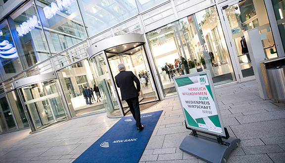 Ein Mann betritt das Foyer der NRW.BANK. Vor der Tür ein Schild: Herzlich willkommen zum MEDIENPREIS WIRTSCHAFT NRW 2021