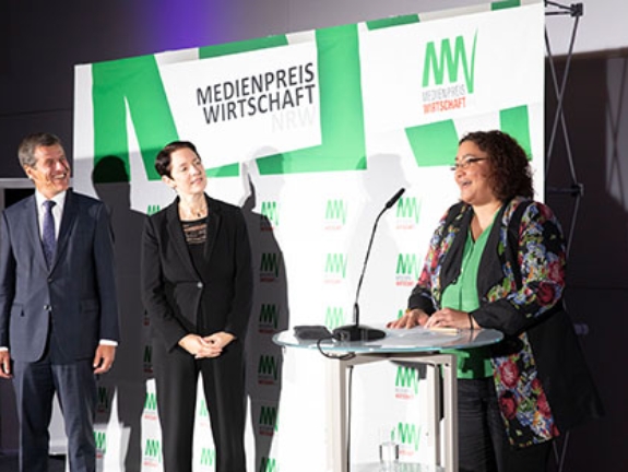 Eckhard Forst, Vorstandsvorsitzender der NRW.BANK, und NRW.Landwirtschaftsministerin Silke Gorißen hören Yasmin Osman zu, die vor einer Logowand eine Laudatio hält.