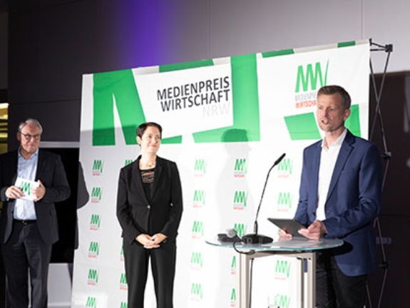 Norbert Tiemann, NRW-Landwirtschaftsministerin Silke Gorißen und Michael Block stehen vor einer Logowand. Michael Block hält eine Laudatio.