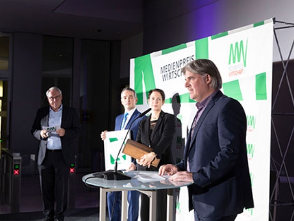 Vor einer Logowand stehen Norbert Tiemann, ein Vertreter der Heidelberger Druckmaschinen AG und NRW-Landwirtschaftsministerin Silke Gorißen, während Ralph Goldmann am Pult steht und eine Laudation hält.