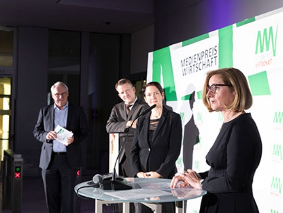 Norbert Tiemann, ein Vertreter der Signal Iduna Gruppe und NRW-Landwirtschaftsministerin Silke Gorißen hören zu, wie Brigitte Koch-Frickenhaus eine Laudatio an einem Pult hält.