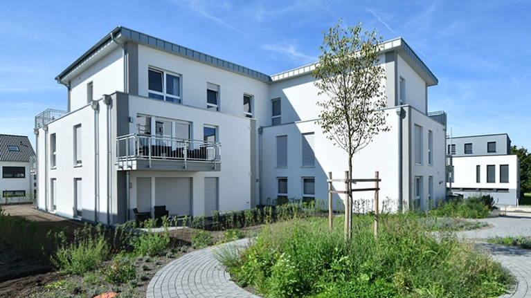 Modernes Wohnhaus mit Balkonen, gepflegter Grünfläche und geschwungenem Weg bei blauem Himmel.