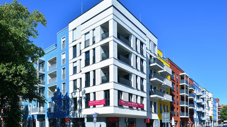 Entlang der Straße reihen sich farbige Fassaden mit Balkonen aneinander, während Menschen vor Läden vorbeigehen.