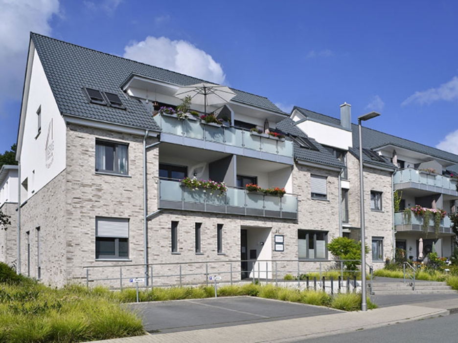 Mehrfamilienhaus mit hellen Klinkern, blumigen Balkonen und gepflegtem Vorgarten bei blauem Himmel.