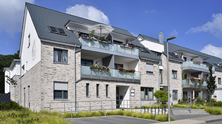 Mehrfamilienhaus mit hellen Klinkern, blumigen Balkonen und gepflegtem Vorgarten bei blauem Himmel.