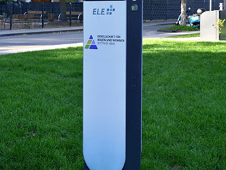 Ladesäule auf grüner Wiese vor modernen Wohnhäusern, umgeben von Herbstbäumen und ruhiger Wohnstraße.