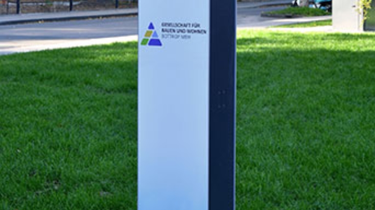 Ladesäule auf grüner Wiese vor modernen Wohnhäusern, umgeben von Herbstbäumen und ruhiger Wohnstraße.