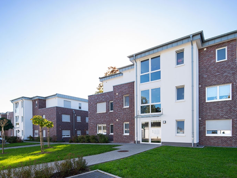 Moderne Mehrfamilienhäuser aus Klinker und Weiß stehen in grüner Anlage, Wege führen durch gepflegte Rasenflächen.