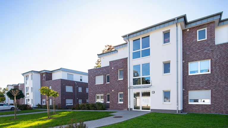 Moderne Mehrfamilienhäuser aus Klinker und Weiß stehen in grüner Anlage, Wege führen durch gepflegte Rasenflächen.