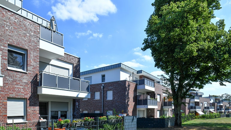 Backsteinanlage mit Balkonen, Gärten und großem Baum in einer sonnigen, ruhigen Nachbarschaft.