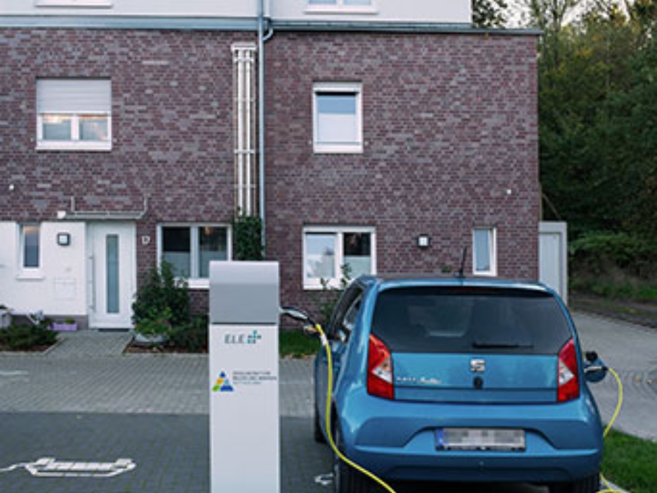 Blaues E-Auto lädt vor einem Backsteinhaus an einer Stromsäule, umgeben von Grün und Abendlicht.