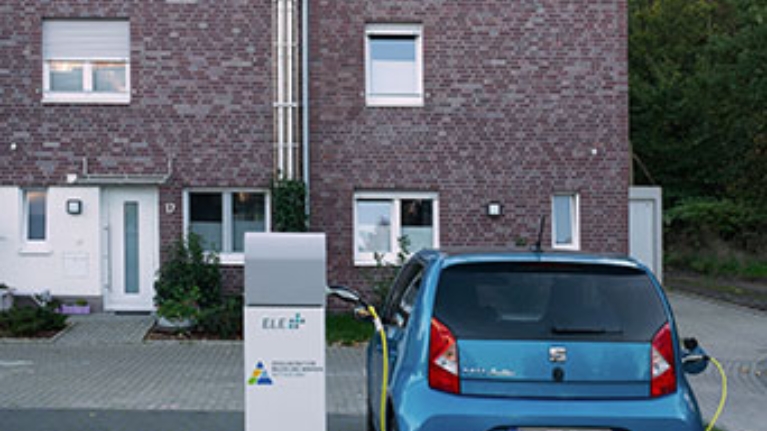 Blaues E-Auto lädt vor einem Backsteinhaus an einer Stromsäule, umgeben von Grün und Abendlicht.