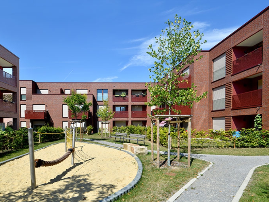 Klinkerwohnanlage mit Balkonen, begrüntem Innenhof und Spielplatz unter blauem Himmel.