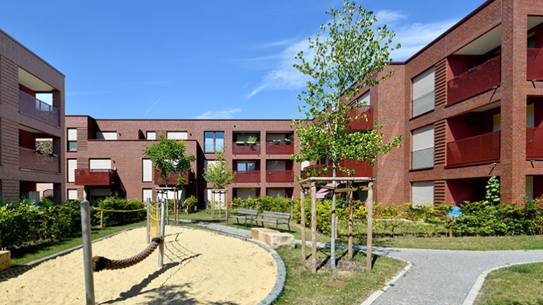 Klinkerwohnanlage mit Balkonen, begrüntem Innenhof und Spielplatz unter blauem Himmel.
