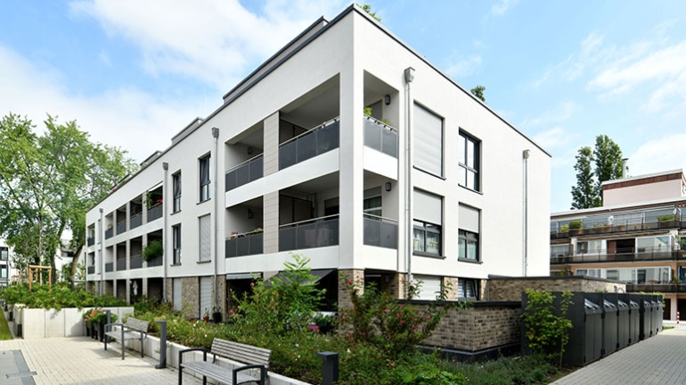 Weiße Wohnanlage mit breiten Balkonen, Sitzbänken und grüner Bepflanzung unter blauem Sommerhimmel.