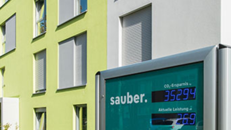 Digitales Display vor grüner Fassade zeigt CO₂-Ersparnis und Leistung des Mieterstroms.
