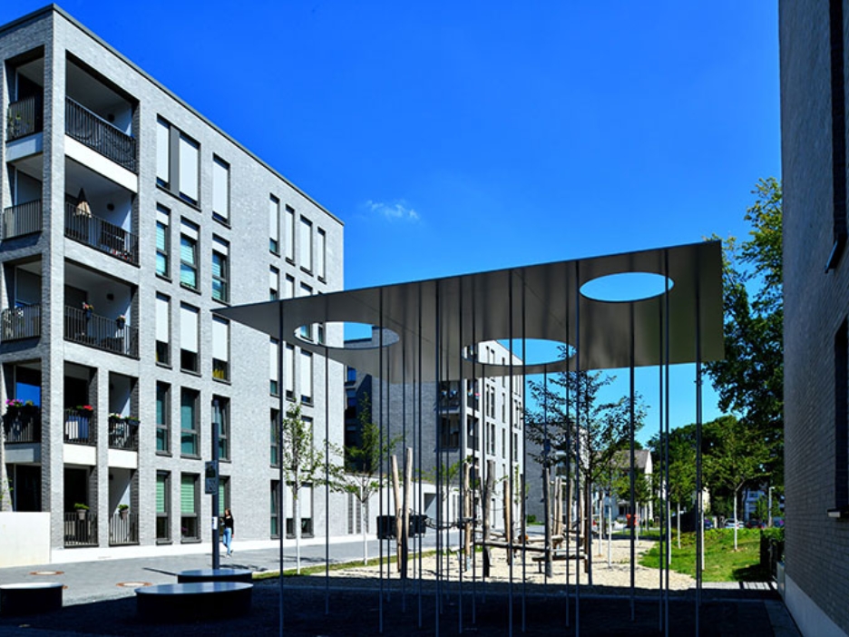 Zwischen modernen Gebäuden liegt ein Platz mit schattenspendender Metallkonstruktion und Holzspielgeräten.