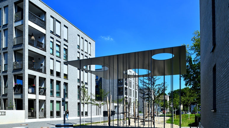Zwischen modernen Gebäuden liegt ein Platz mit schattenspendender Metallkonstruktion und Holzspielgeräten.