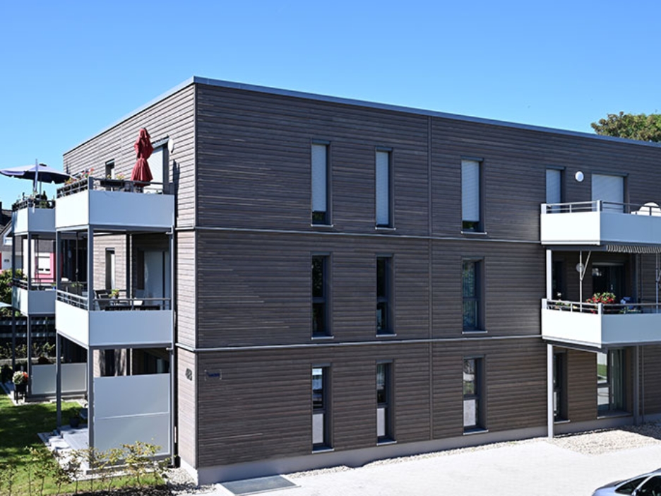 Modernes Wohnhaus mit Holzfassade und hellen Balkonen in ruhiger Nachbarschaft bei blauem Himmel.