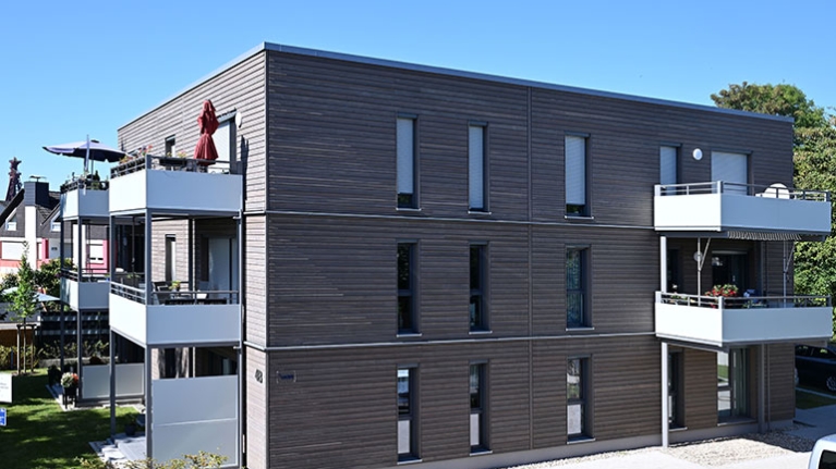 Modernes Wohnhaus mit Holzfassade und hellen Balkonen in ruhiger Nachbarschaft bei blauem Himmel.