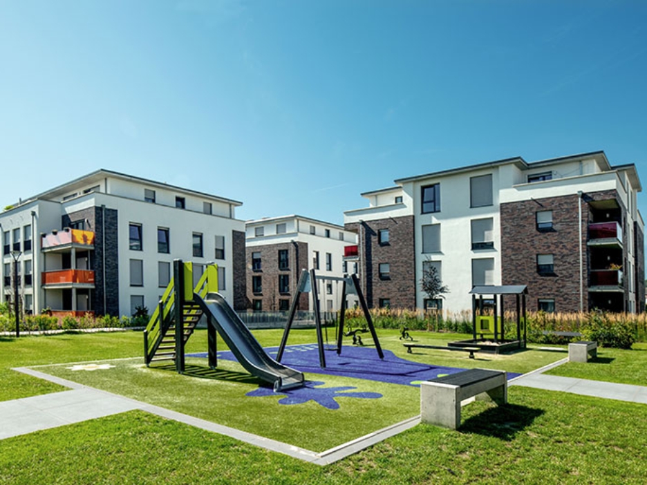 Helle Wohngebäude umrahmen einen großen grünen Hof mit Spielplatz, darüber erstreckt sich ein klarer blauer Himmel.