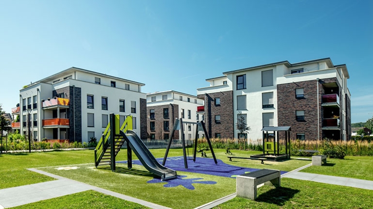 Helle Wohngebäude umrahmen einen großen grünen Hof mit Spielplatz, darüber erstreckt sich ein klarer blauer Himmel.