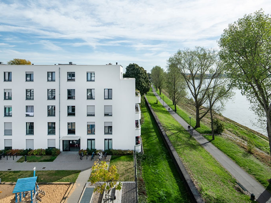 Weißes Wohnhaus am Rhein mit Grünanlage, Uferweg und großem Baumbestand bei leichtem Himmel.