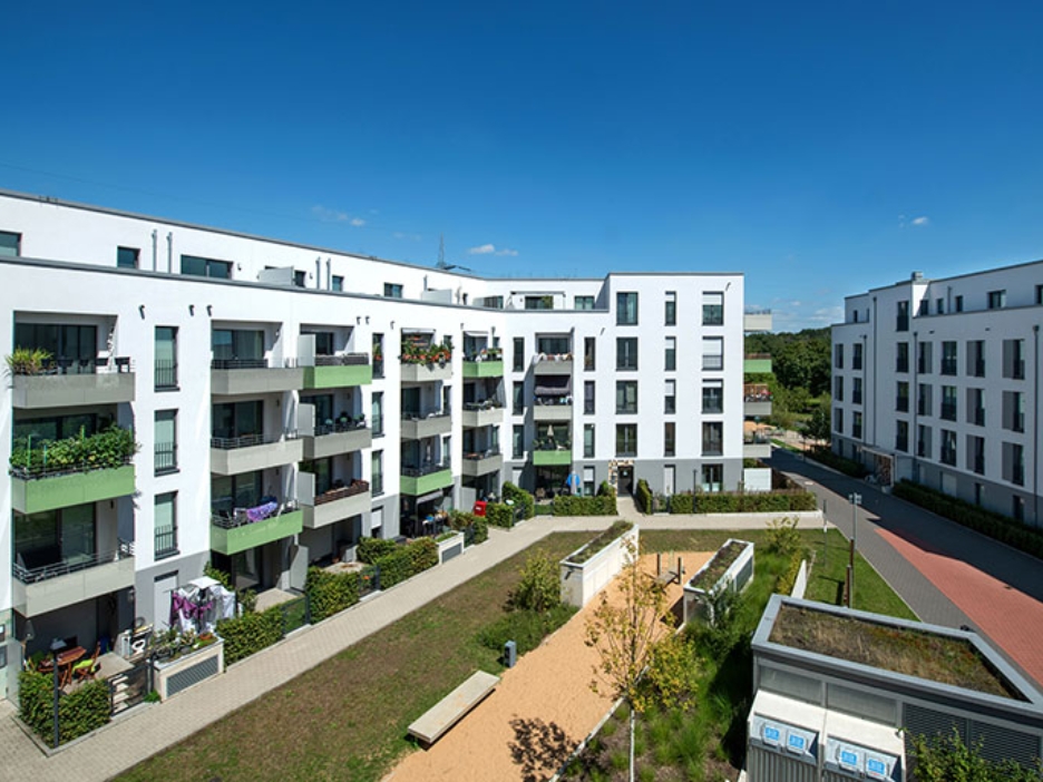 Helle Wohnhäuser mit grünen Balkonen rahmen einen begrünten Innenhof, in dem Wege und Beete eine ruhige Nachbarschaftsatmosphäre schaffen.