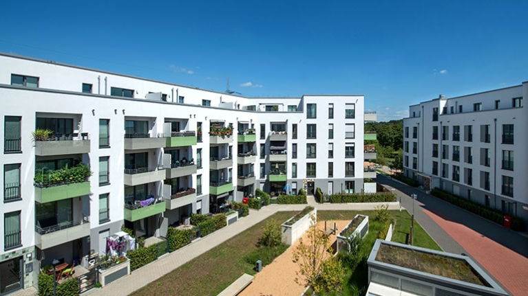 Helle Wohnhäuser mit grünen Balkonen rahmen einen begrünten Innenhof, in dem Wege und Beete eine ruhige Nachbarschaftsatmosphäre schaffen.